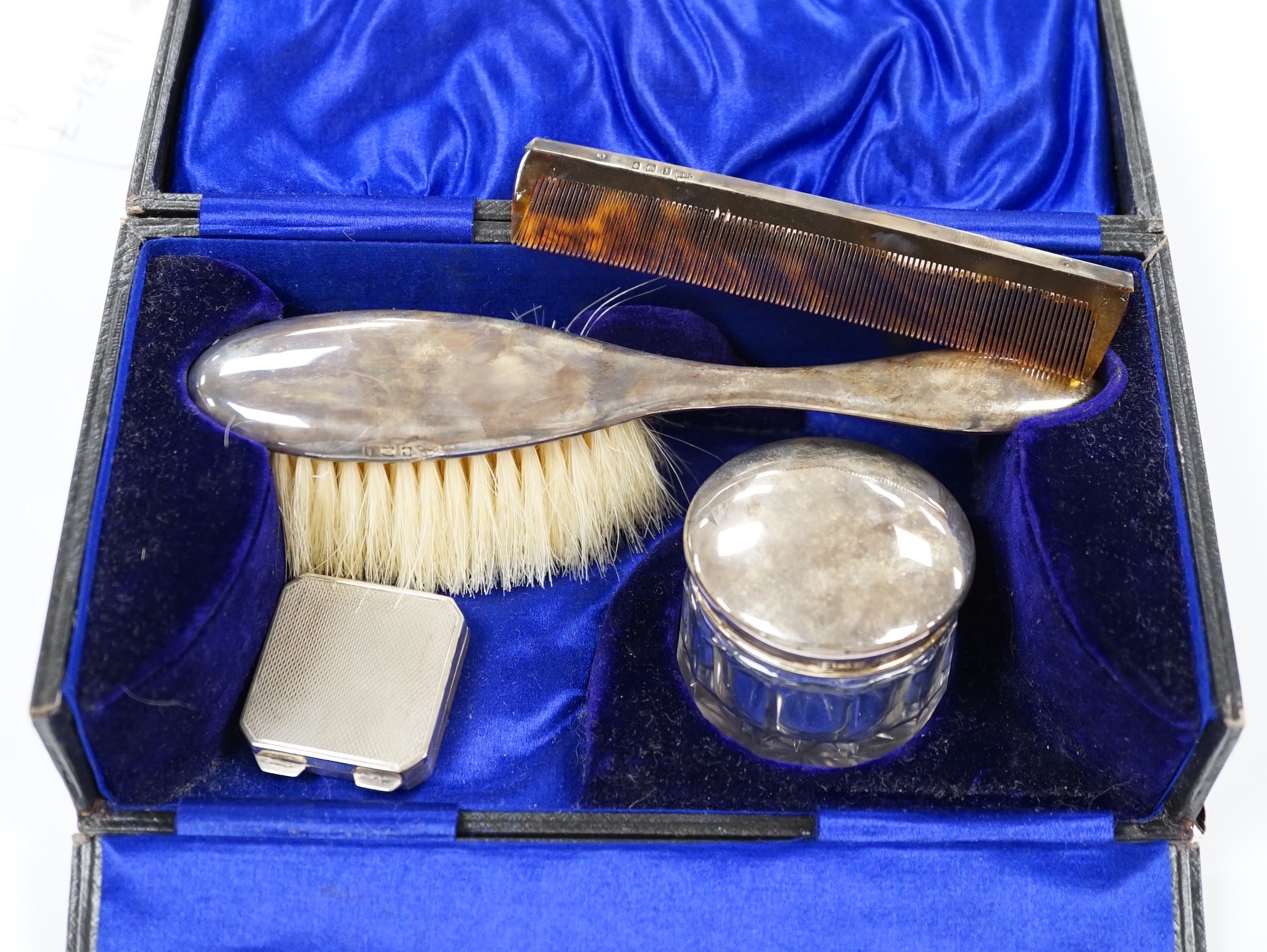 A George V engine turned silver pill box, 34mm and a cased silver mounted three piece toilet set.